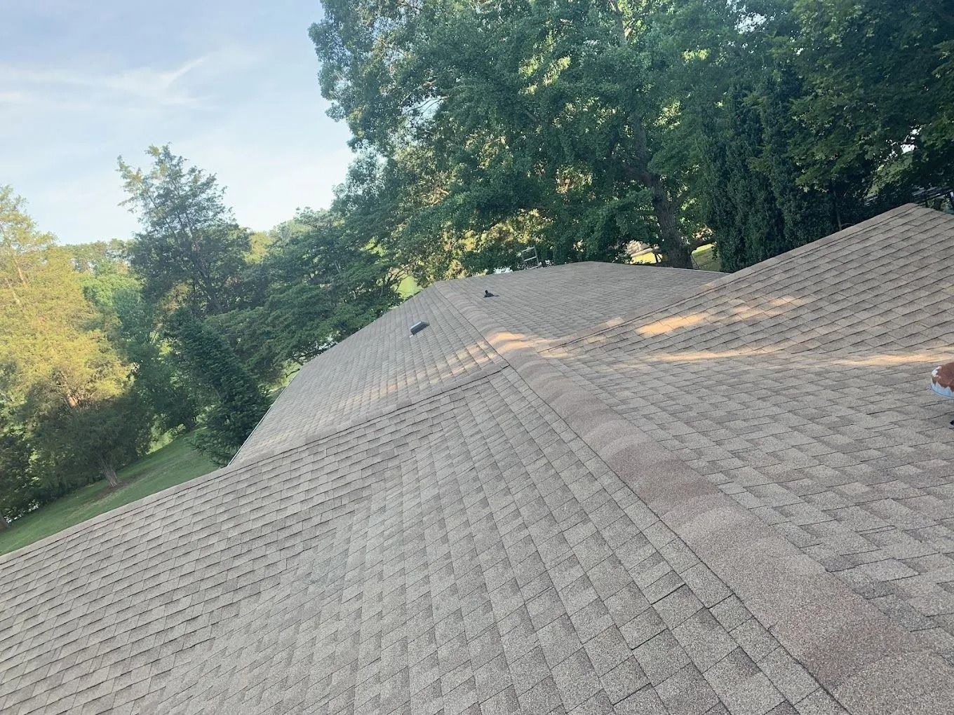 A view looking down over the brown shingled roof of a house, surrounded by lush green trees under a bright sky.