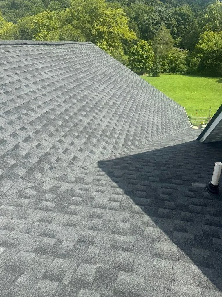A high-angle view of a grey shingled roof meeting at a valley, with trees and a green lawn visible in the background.
