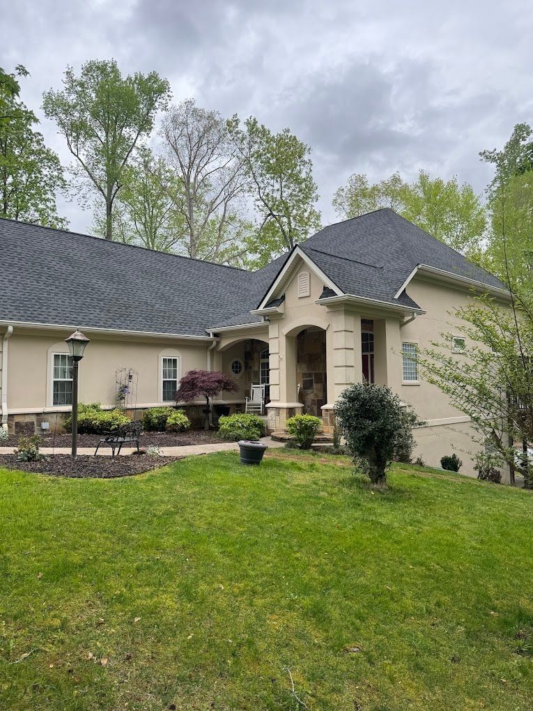 A beige house with a dark shingled roof, set on a grassy lot with trees under a cloudy sky.