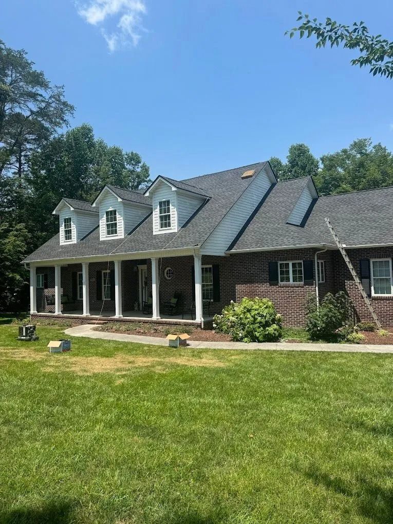 A two-story brick house with a covered front porch, three gabled dormers, and a gray shingle roof in a grassy yard.