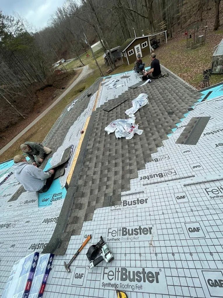 Three workers installing gray asphalt roof shingles onto a sloped roof covered in protective felt underlayment.