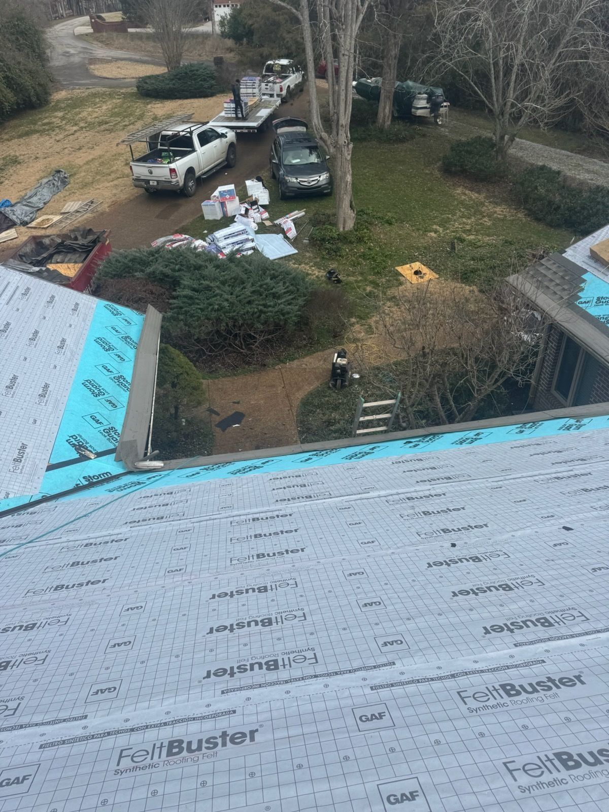 An elevated view of a house roof undergoing construction, with work trucks and materials scattered on the lawn below.