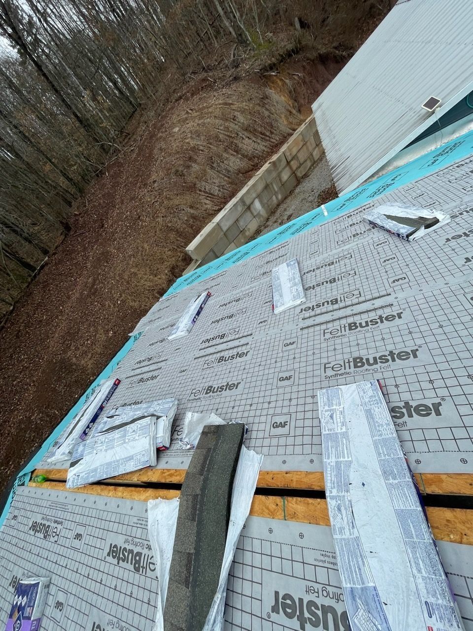 A roof under construction, featuring gray synthetic underlayment sheets laid over wood decking near a wooded area.