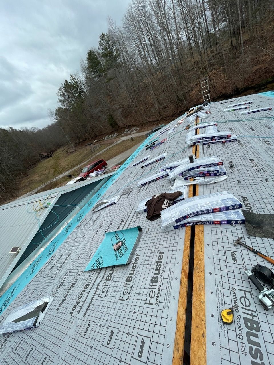 A sloped roof under construction, covered in gray felt paper with shingles stacked along the ridge, outdoors.