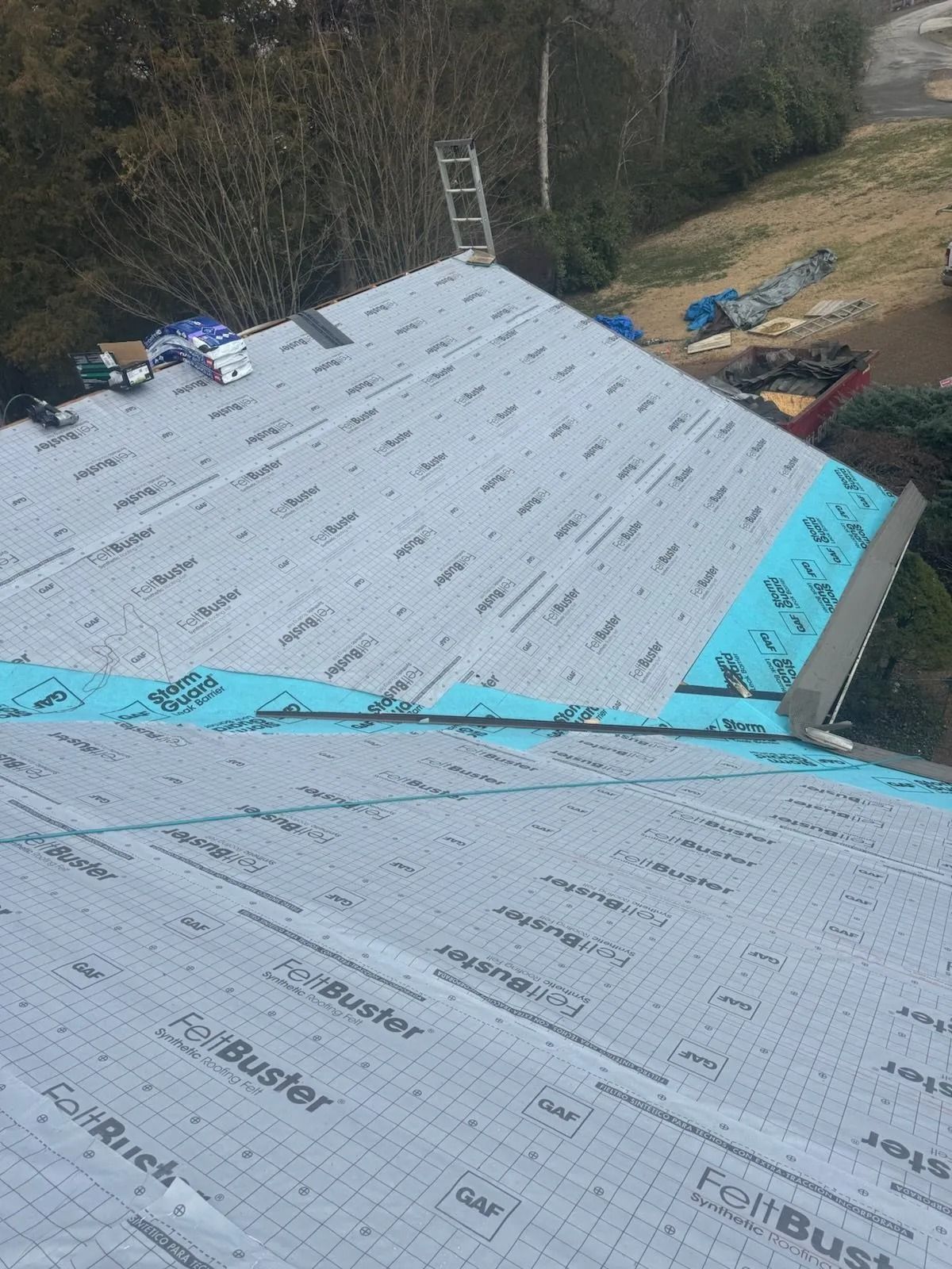 A house roof under construction, covered in gray FeltBuster underlayment and blue waterproof barrier along the edges.