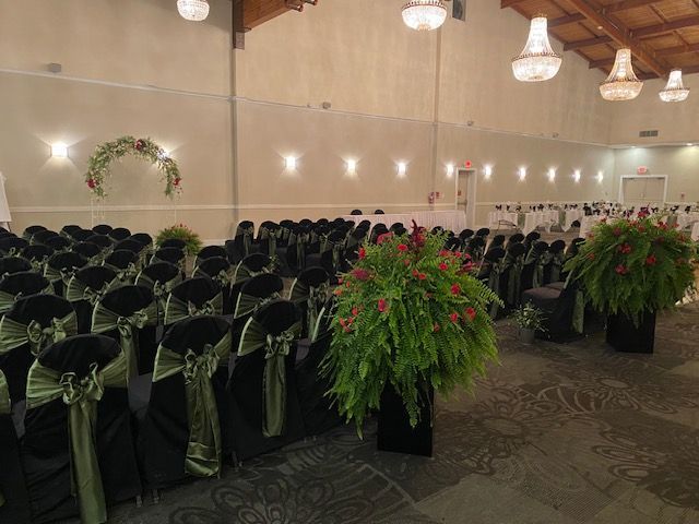 An empty wedding reception hall with black chairs decorated with green sashes, fern arrangements, and a floral arch.