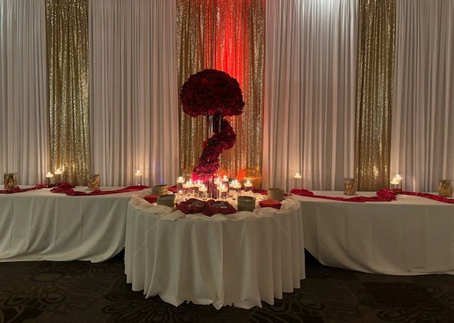 Elegant event decor with red floral centerpiece, candles on a white table, and gold and white drapery backdrop.