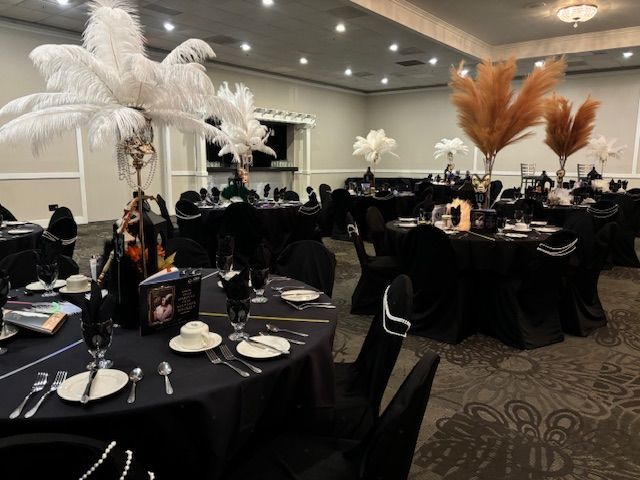 Round tables with black tablecloths, decorated with white and tan feather centerpieces, set for a formal event.