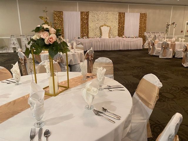 Wedding reception hall, tables set with gold and white decor, a decorated stage, and floral arrangements.