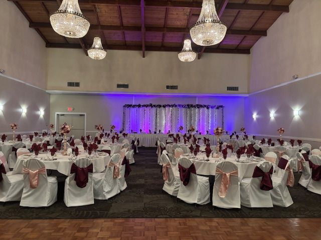 Wedding reception hall with decorated tables and a stage; white and burgundy accents with crystal chandeliers.