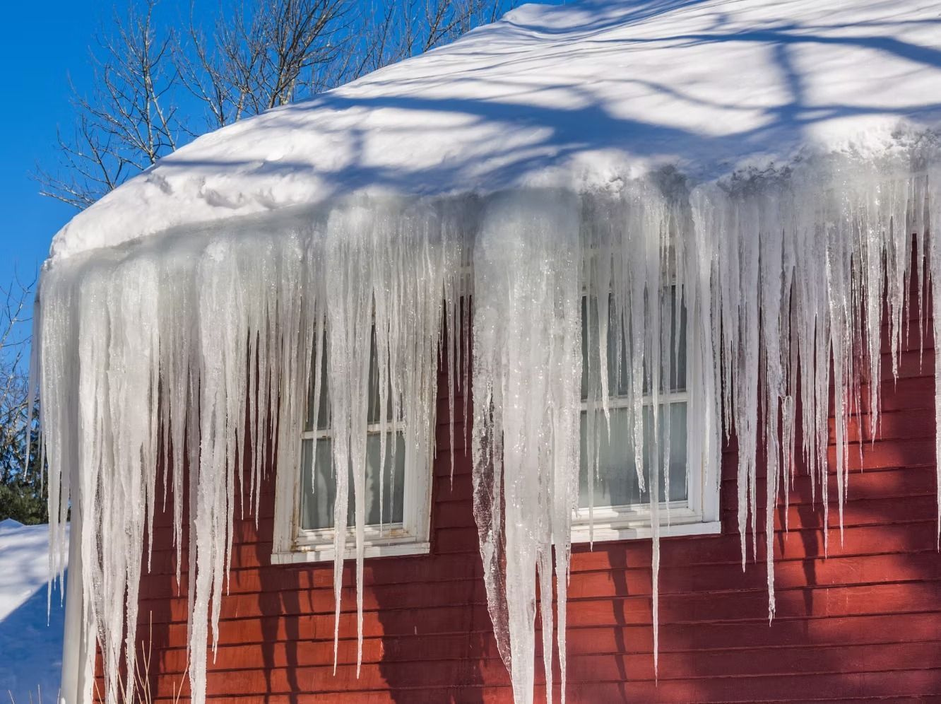 Large icicles hang from the snow-covered roof of a red building on a sunny day.