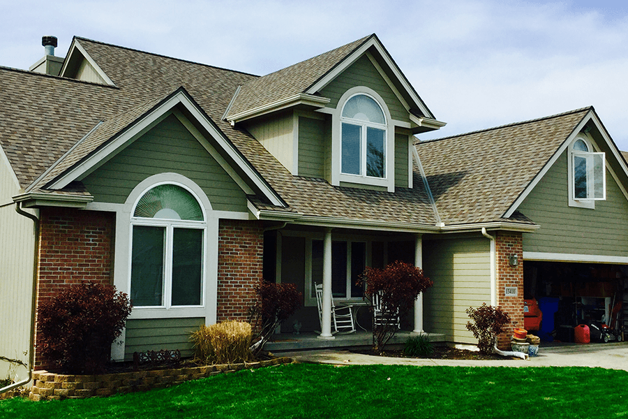 A two-story suburban home with green siding, brick accents, arched windows, and a shingled roof under a blue sky.