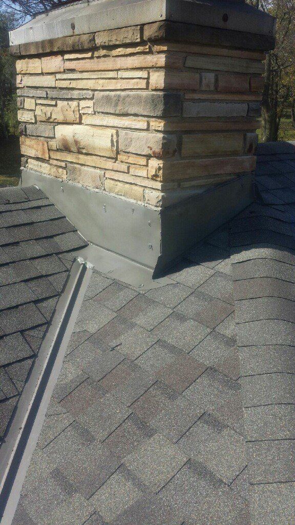 A stone chimney on a residential roof, featuring a metal flashing seal where the stone meets the grey asphalt shingles.