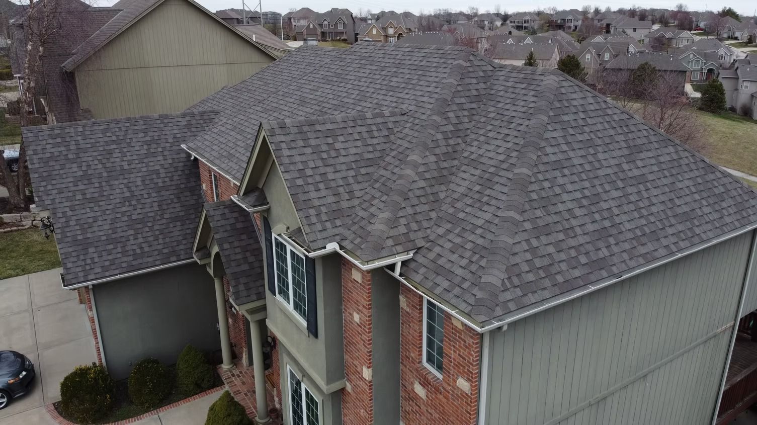 An aerial view of a multi-story home with a grey shingled roof, brick and beige siding, situated in a suburban neighborhood.