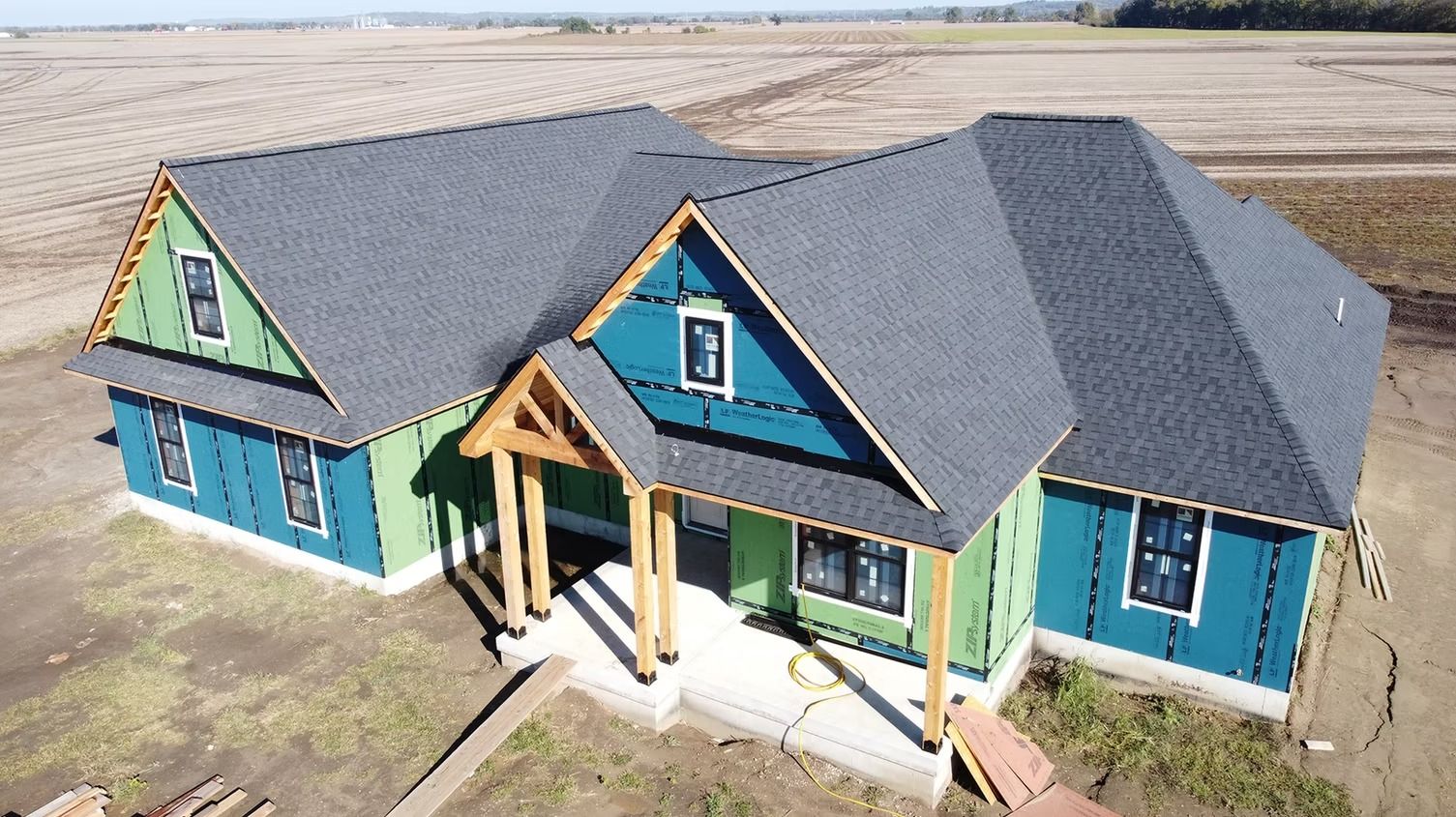 An aerial view of a single-story house under construction with green and blue exterior sheathing and a dark shingled roof.