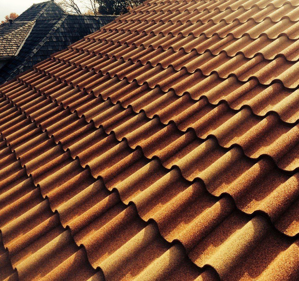 A view looking up at a textured, terracotta-colored tiled roof, with a darker shingled roof visible in the background.