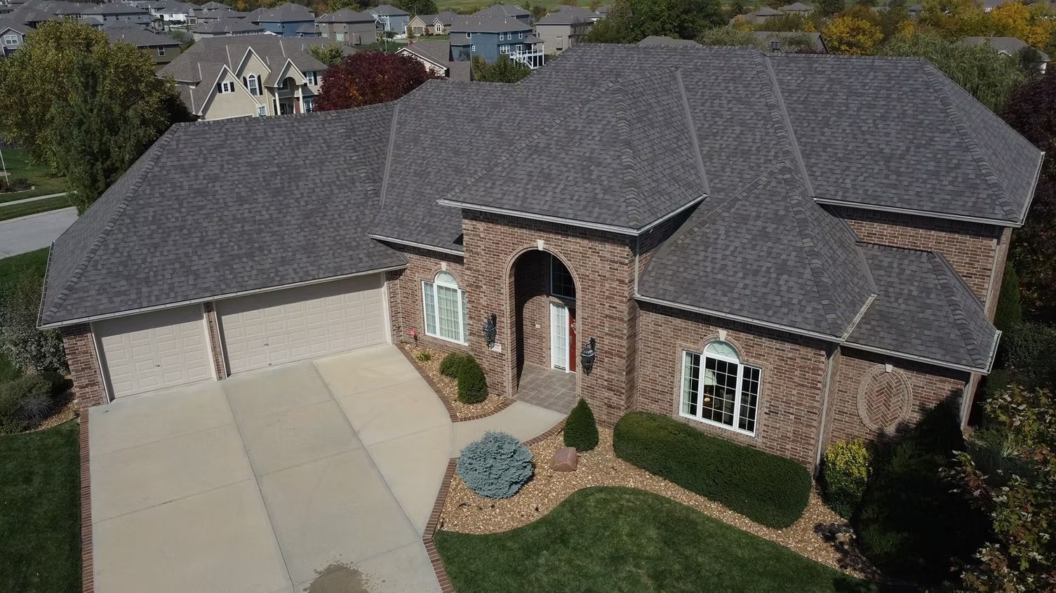 An aerial view of a brick house with a dark gray shingled roof, a paved driveway, and a front yard with manicured bushes.