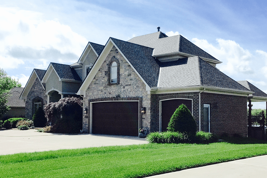 A large, multi-gabled house with a stone and brick exterior, a dark multi-car garage, and a lush green lawn.