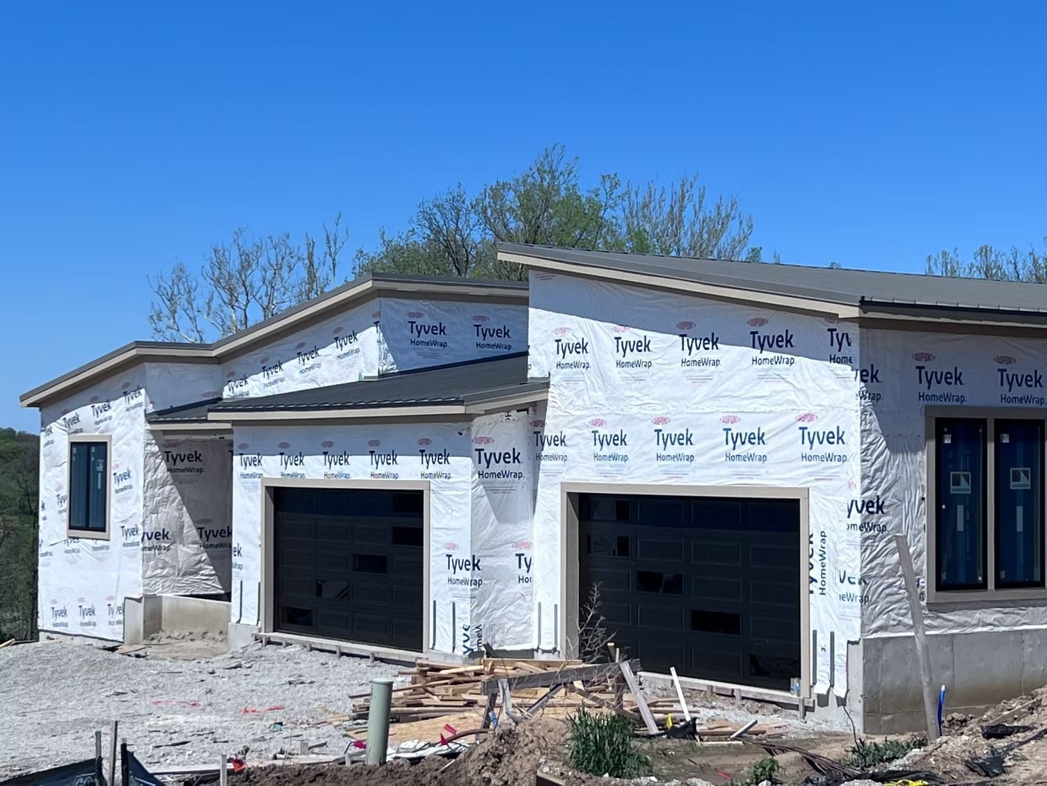 A new single-story house under construction, covered in Tyvek wrap, with two garage doors and visible window frames.
