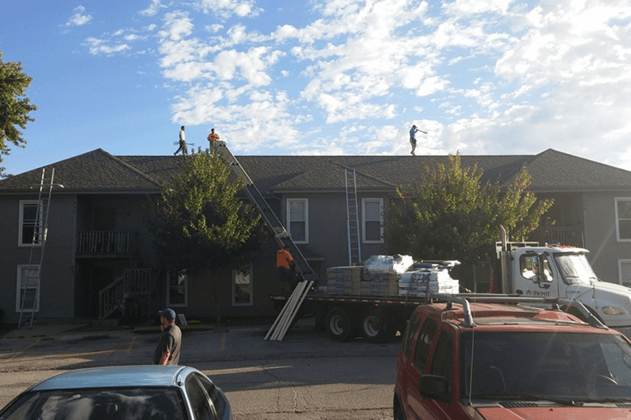 Roofing crew working on top of a gray two-story building with a flatbed truck delivering supplies nearby.
