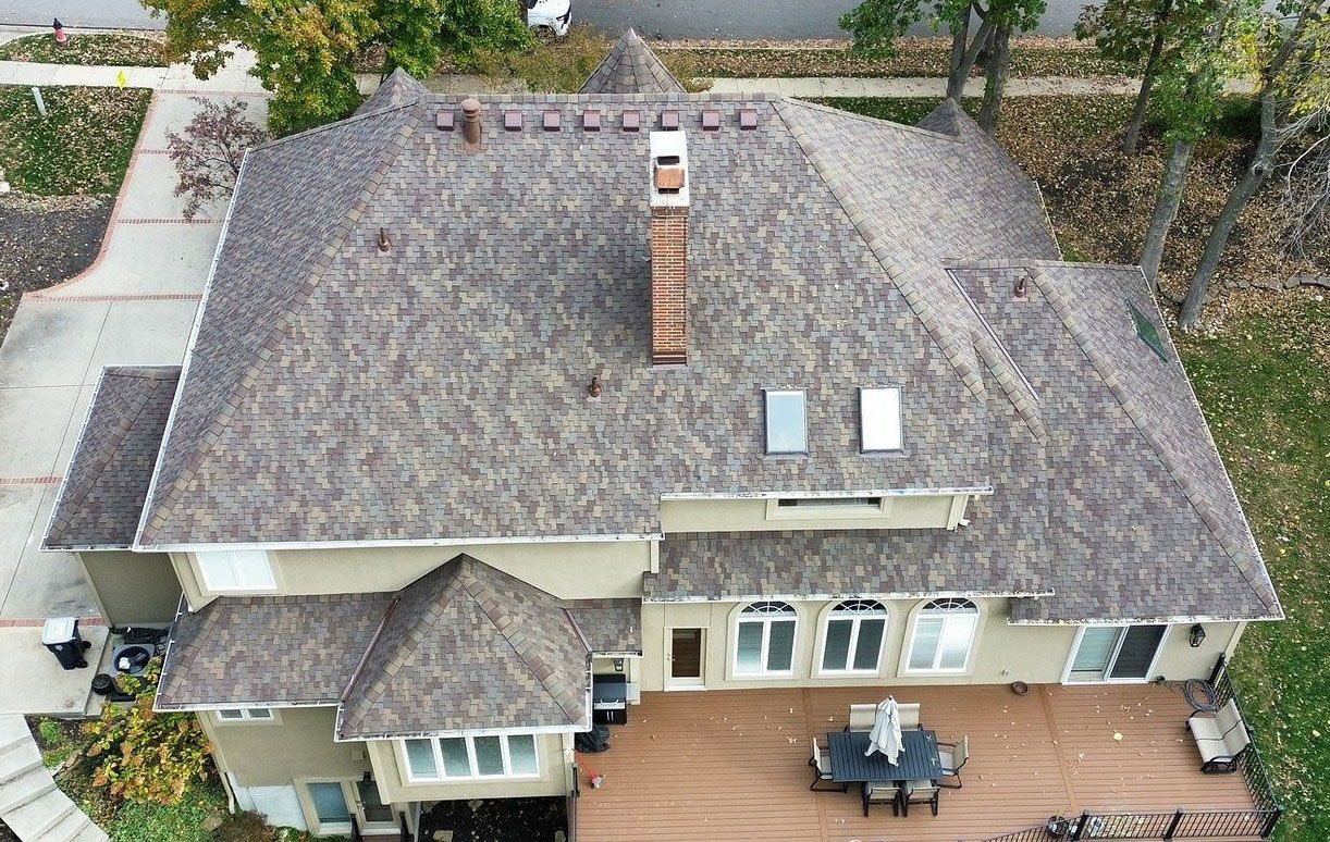An aerial view of a tan two-story house with a shingled roof, a brick chimney, and a wooden deck with patio furniture.