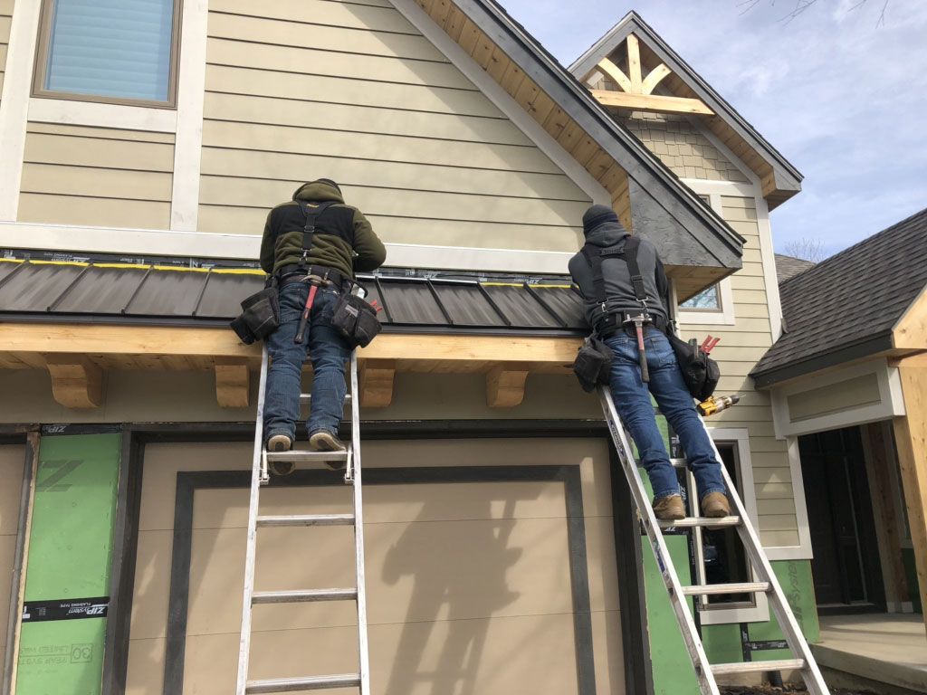 Two workers on ladders install metal roofing over a garage door on a tan house with wooden trim under construction.