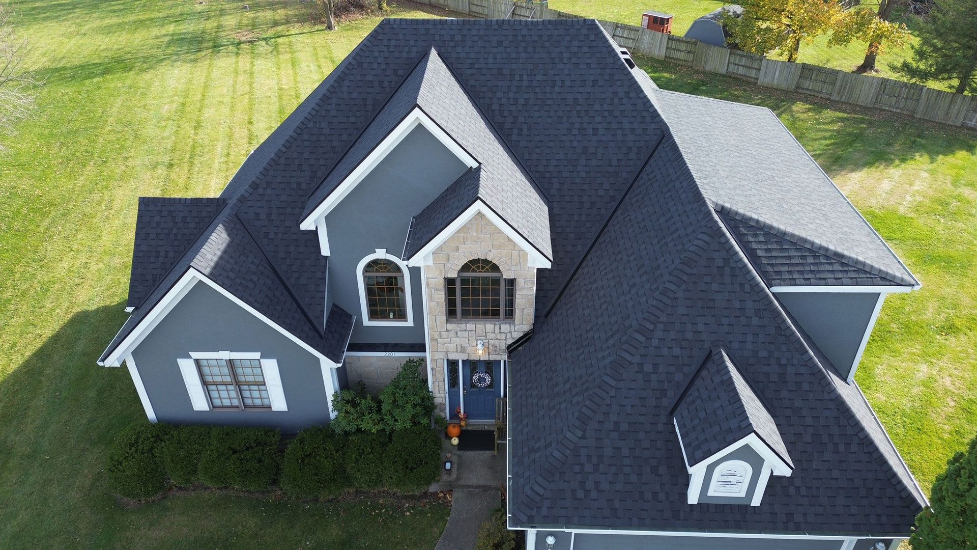 An aerial view of a gray two-story house with a black roof, stone accents, and a blue front door, surrounded by lawn.