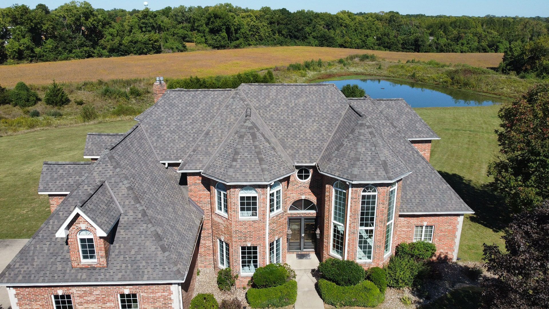 An aerial view of a large, two-story brick house with a gray shingled roof, set in a rural field next to a pond.