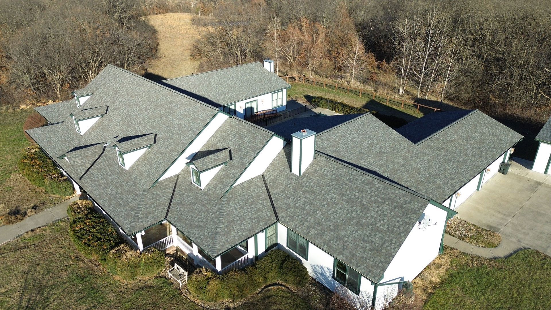 An aerial view of a white, multi-gabled house with a grey shingled roof, surrounded by a lawn and dense trees.