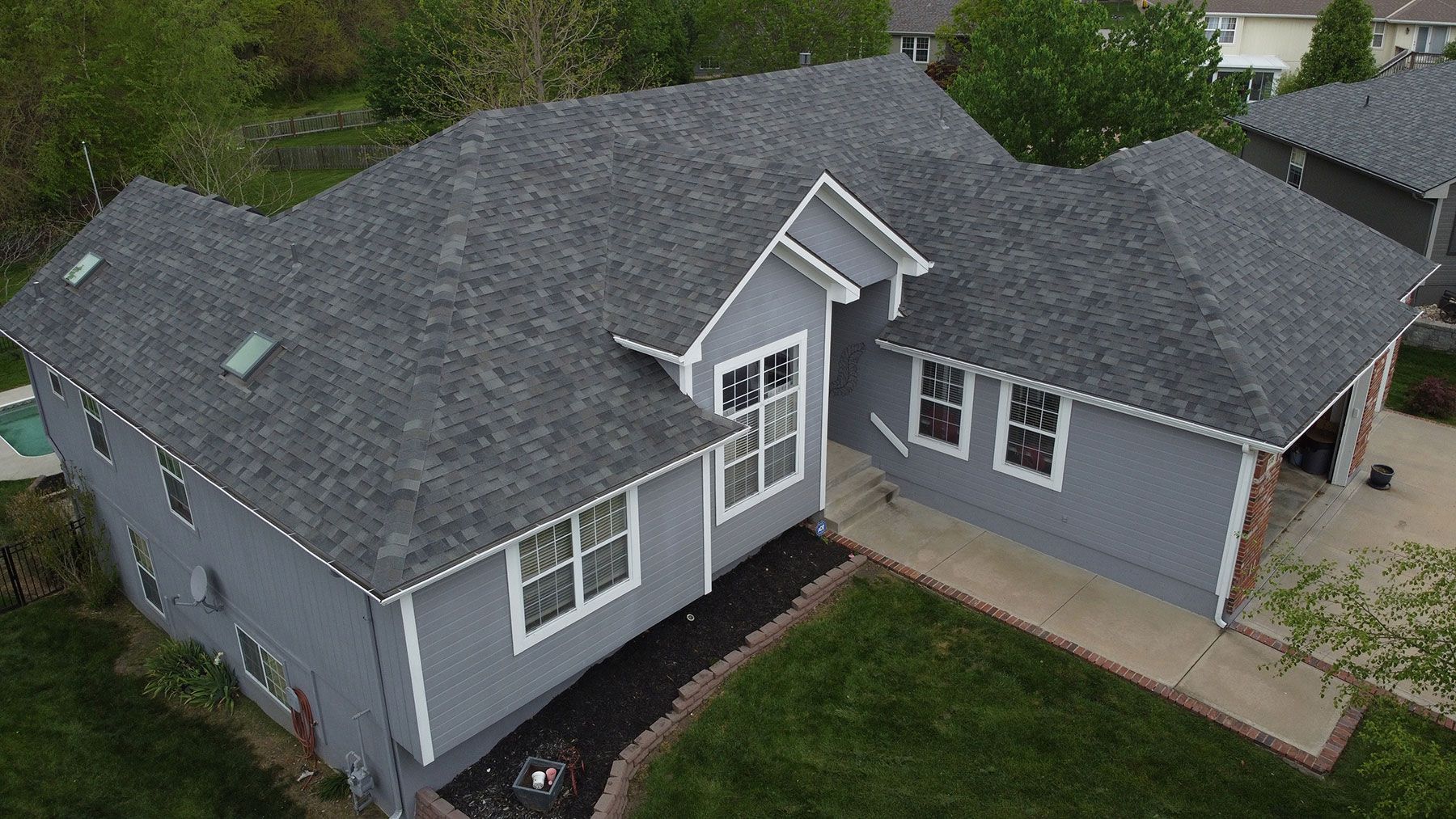 An aerial view of a gray, single-story suburban house with a dark shingled roof, lawn, and concrete driveway.