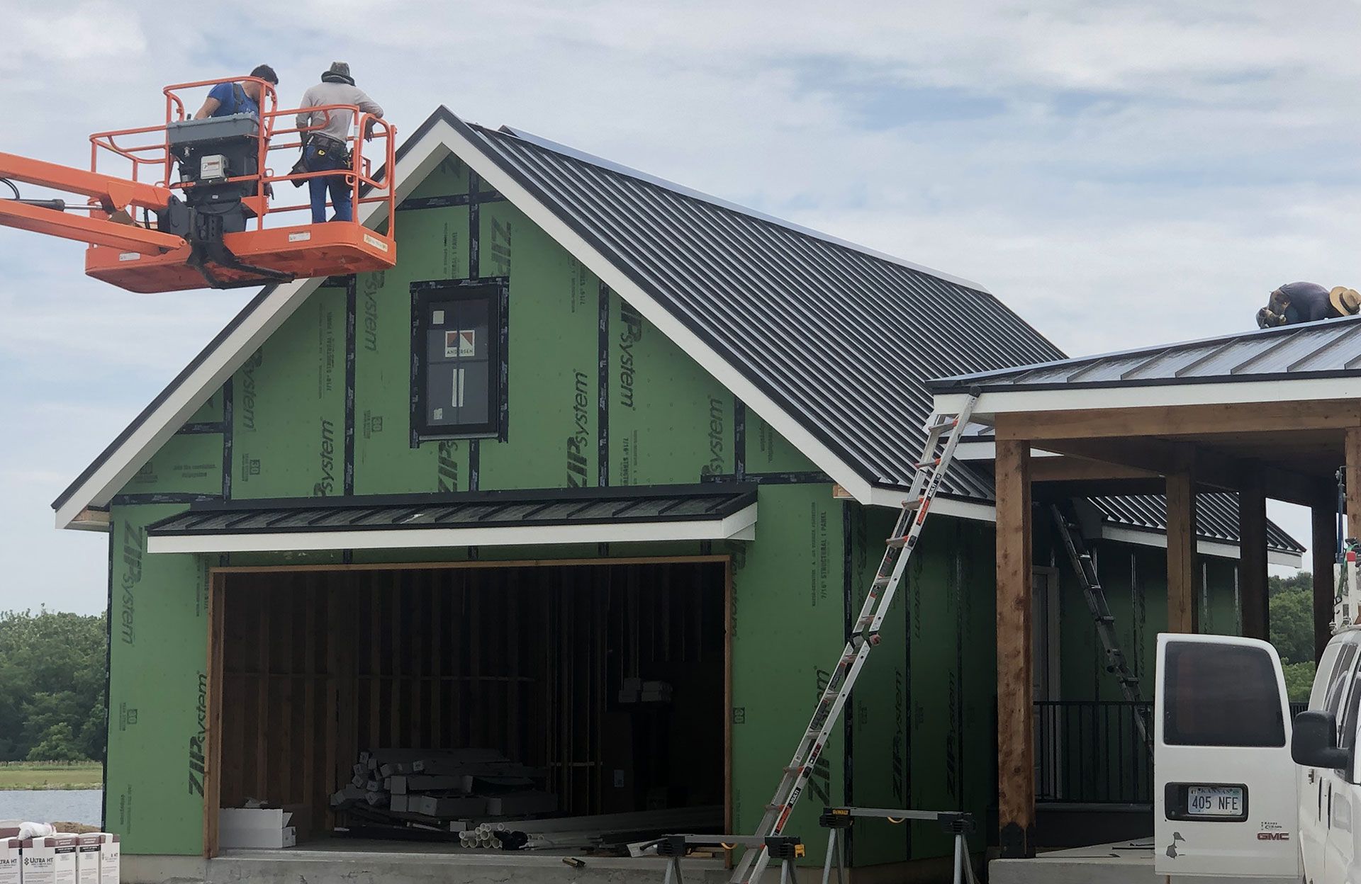 Construction workers on an aerial lift and ladder install a black metal roof on a new house with green exterior sheathing.