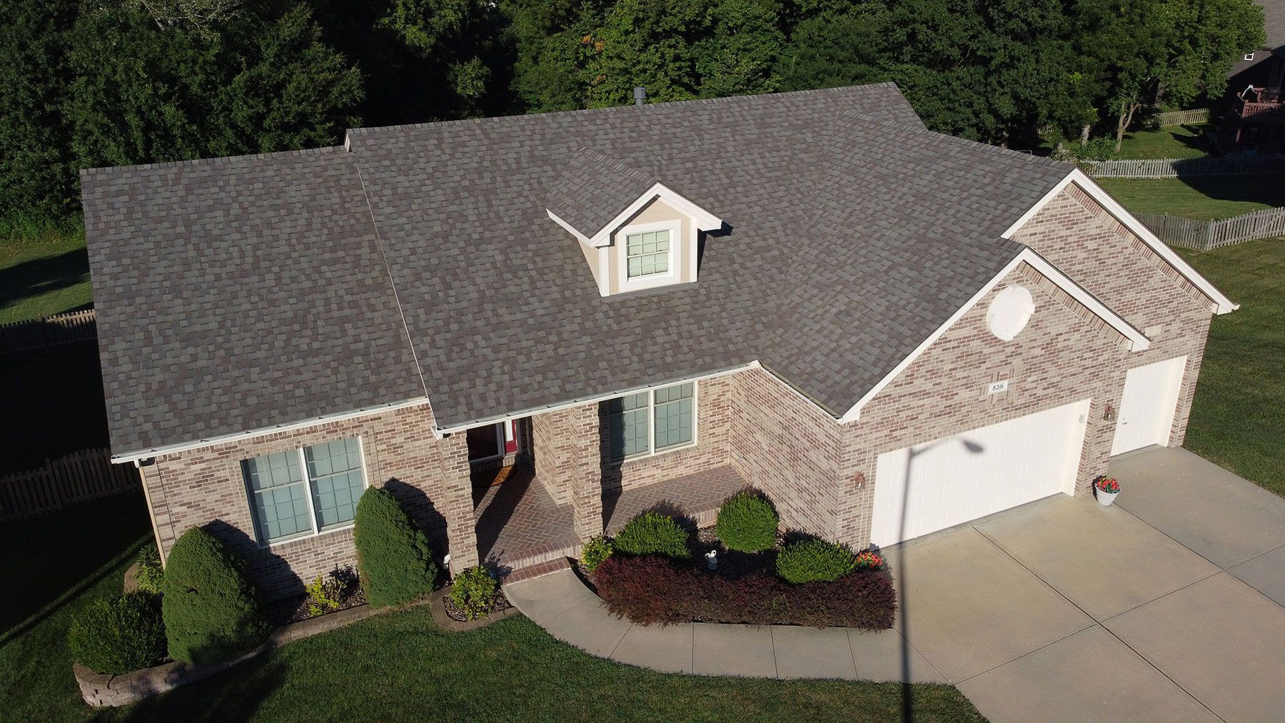 An aerial view of a tan brick suburban house with a gray shingled roof, front landscaping, and a two-car garage.