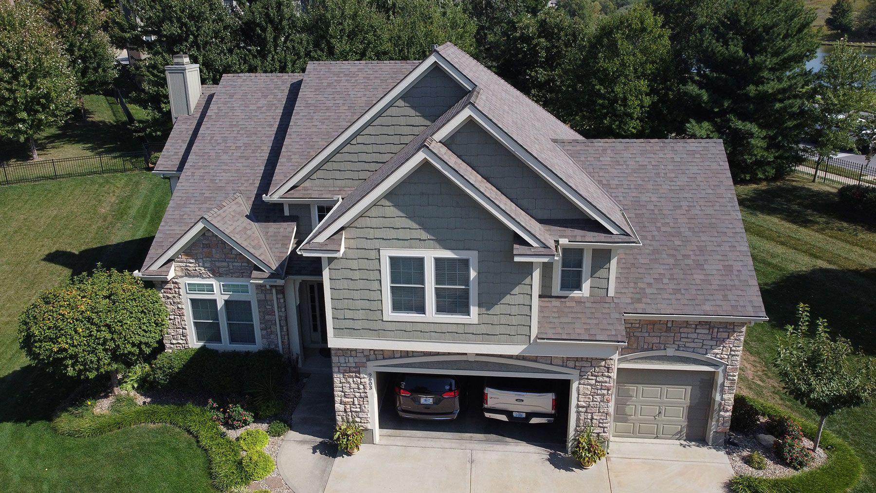 A high-angle view of a multi-story house with a grey shingled roof, light-colored siding, stone accents, and a garage.