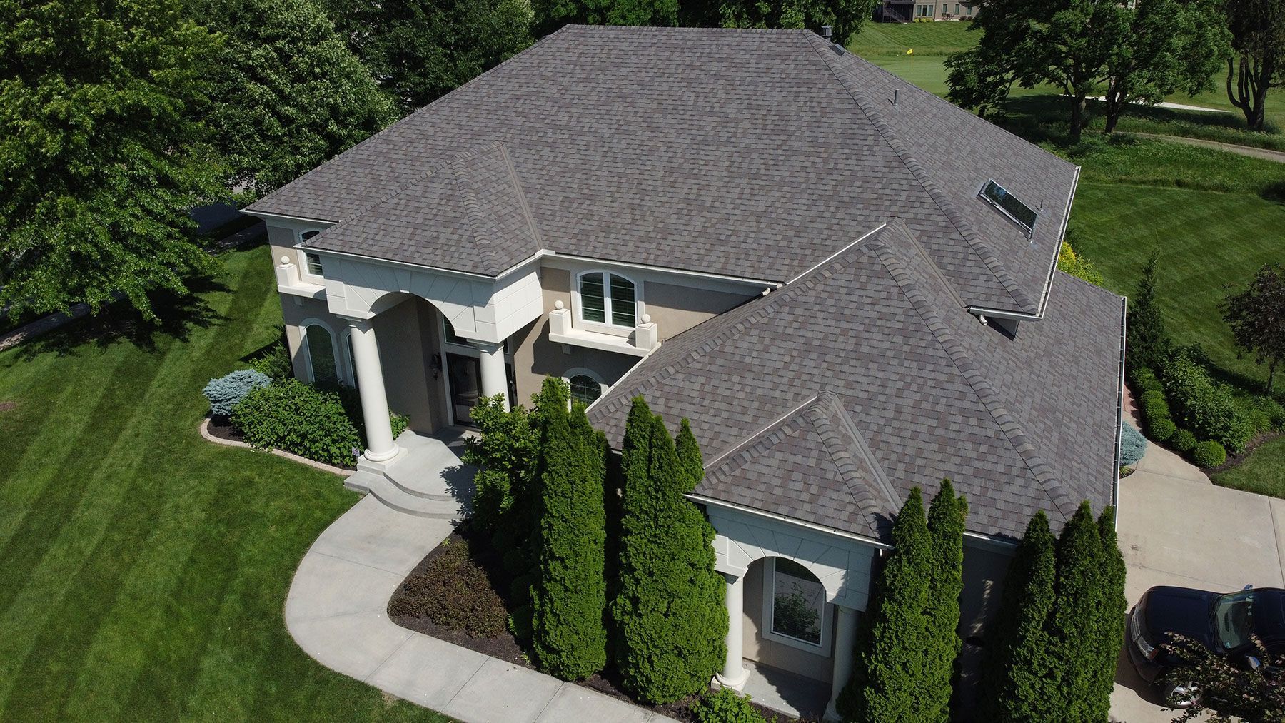 Aerial view of a suburban home with a gray shingled roof, arched entrance, and neatly landscaped lawn.