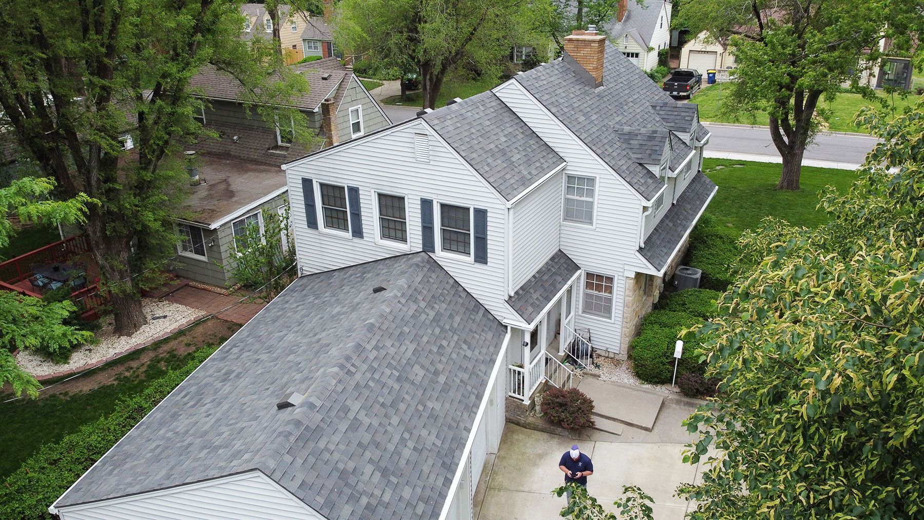 An aerial view of a white, two-story suburban house with a gray shingled roof, surrounded by trees and a paved driveway.