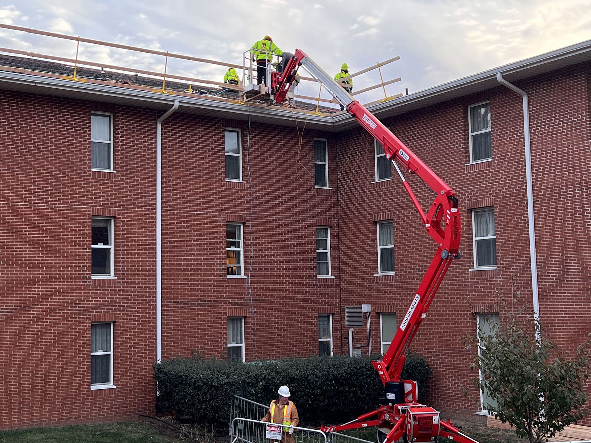 Construction workers in high-visibility gear work on a building roof using a red boom lift in a red brick courtyard.