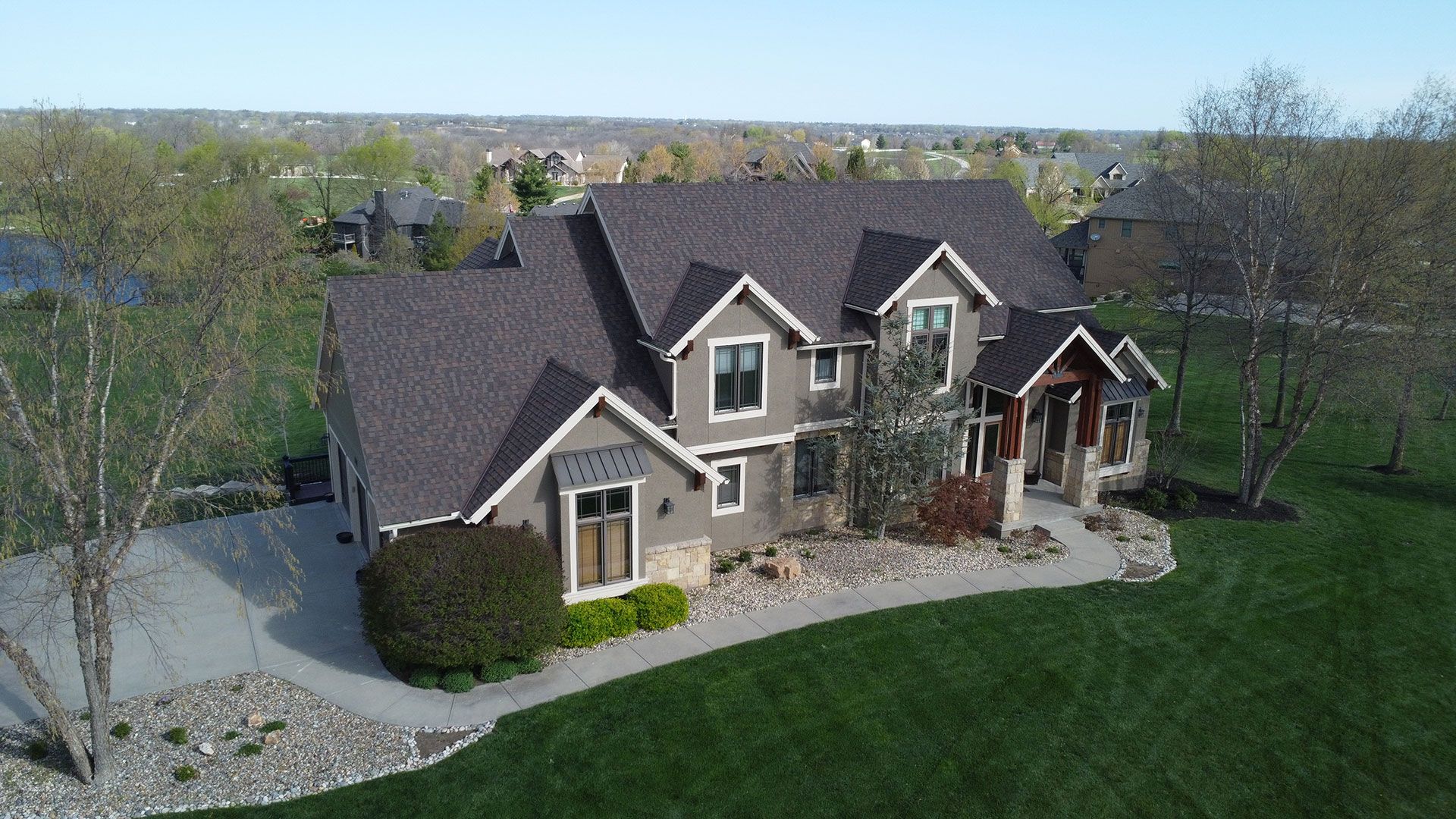 An aerial view of a large, two-story house with dark shingles and tan siding, surrounded by a green lawn and trees.