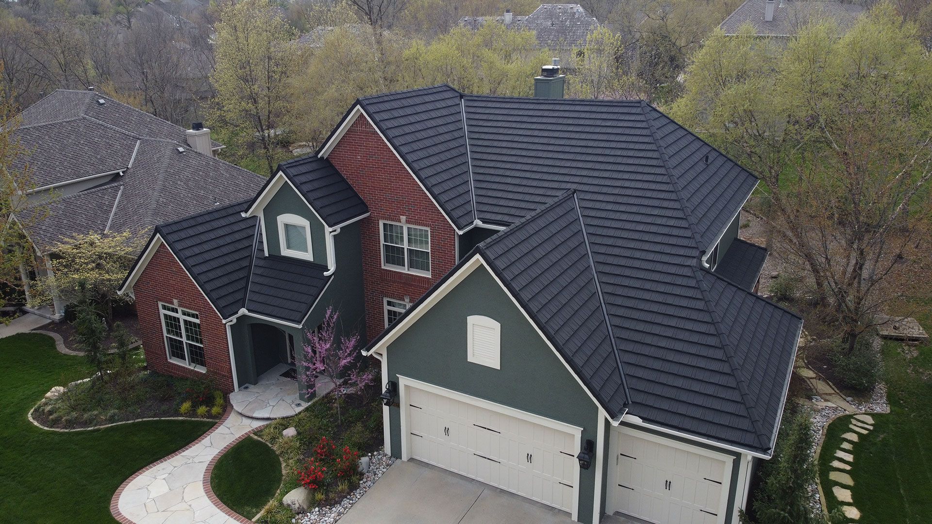 An aerial view of a two-story house with a red and green exterior and a dark shingled roof, surrounded by green trees.