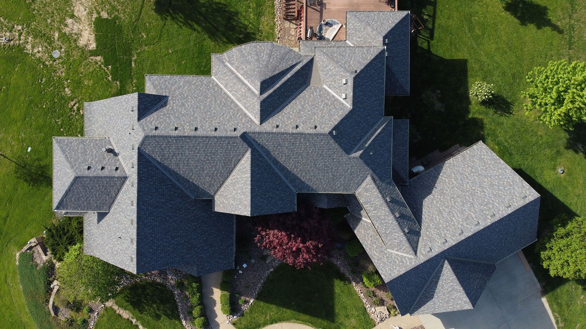Aerial view of a complex, gray shingled residential roof surrounded by a green lawn and trees.