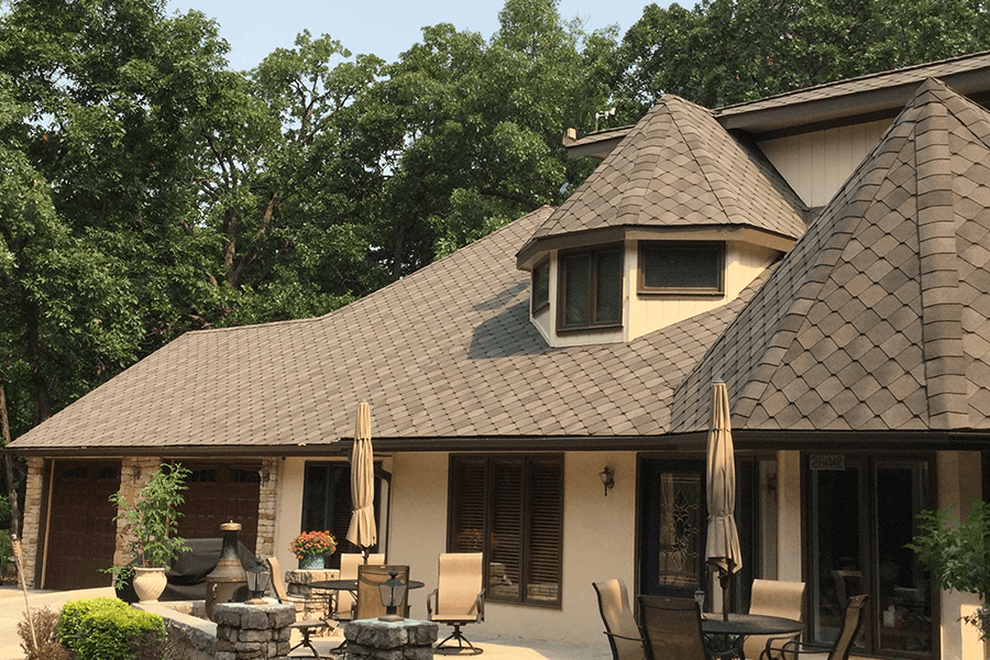 A patio area outside a cream-colored house with shingled roofing, surrounded by green trees on a sunny day.
