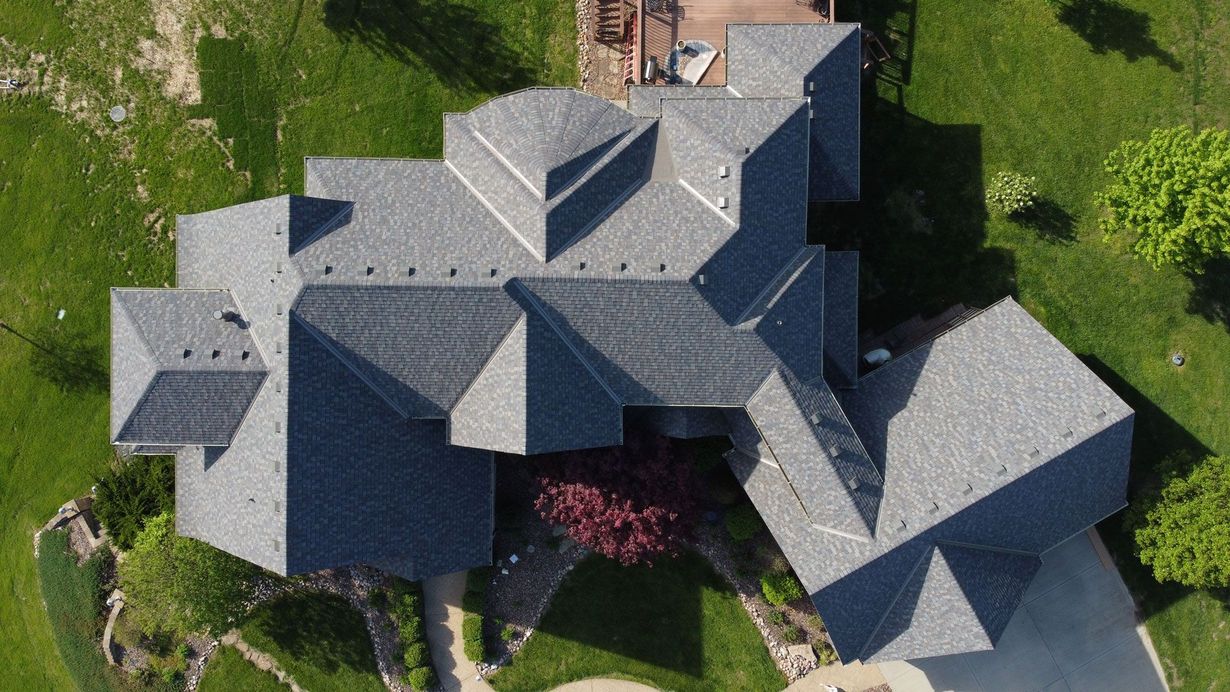 Top-down aerial view of a house with a complex, shingled roof, surrounded by a green lawn and trees.