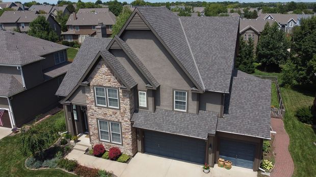 An aerial view of a two-story house with a stone facade, gray exterior, and a dark shingled roof in a suburban neighborhood.