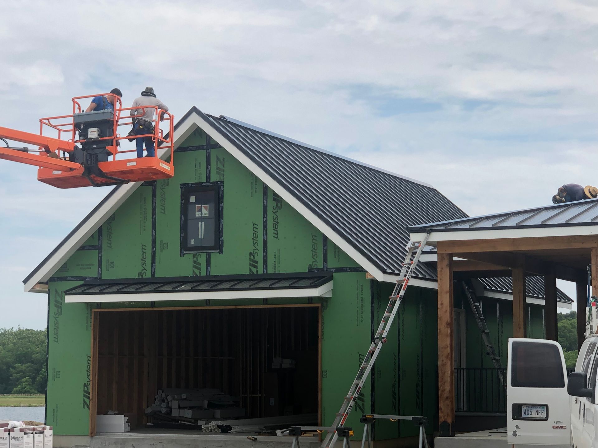 Construction workers on a lift install a metal roof on a green-sheathed, wood-framed building under a bright sky.