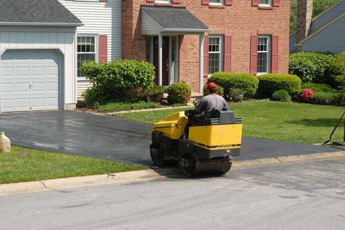 Person riding a yellow lawn mower on a suburban street near a house and driveway