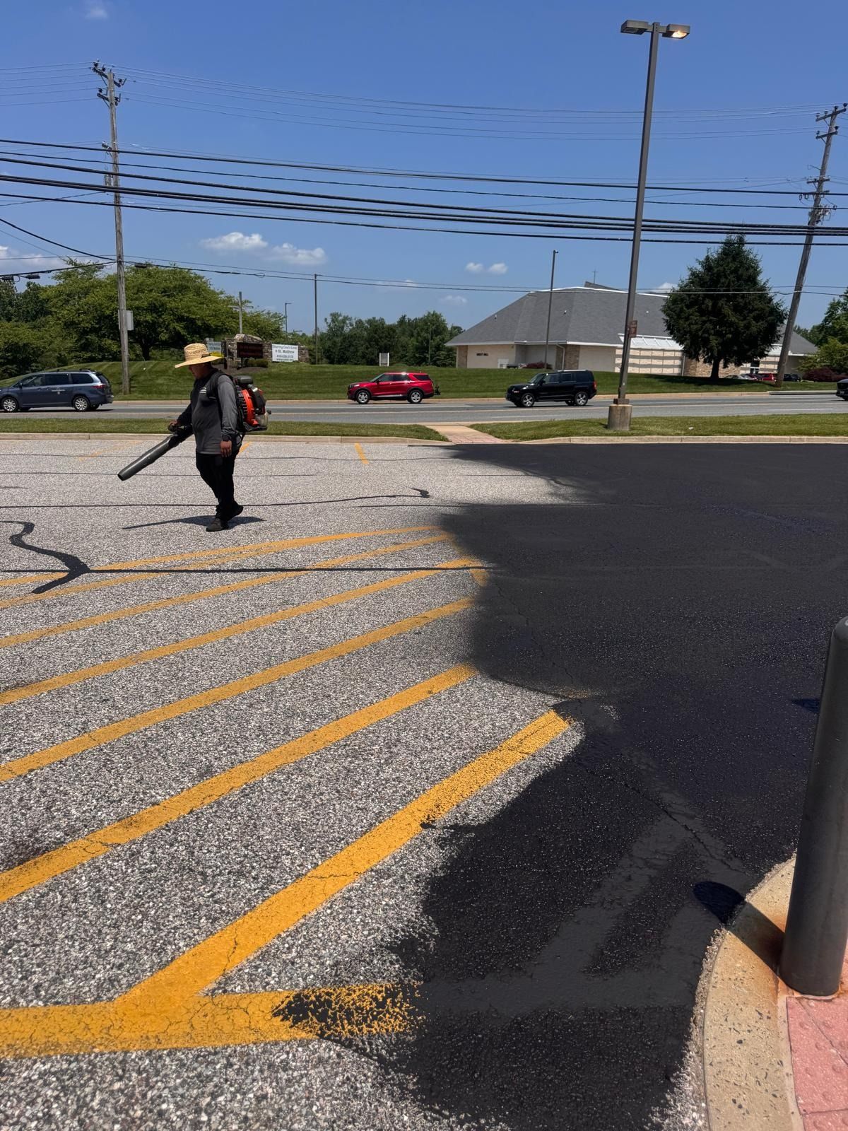 Person crossing a parking lot with yellow stripes near a road, carrying a bag under a clear blue sky.