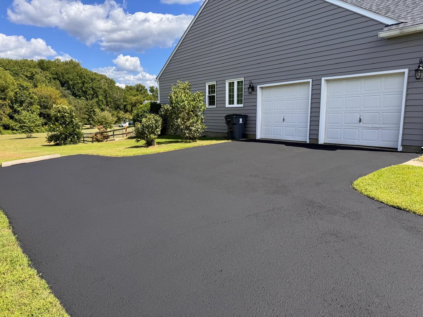 Freshly paved black driveway beside a gray house with two white garage doors and a grassy yard under a blue sky