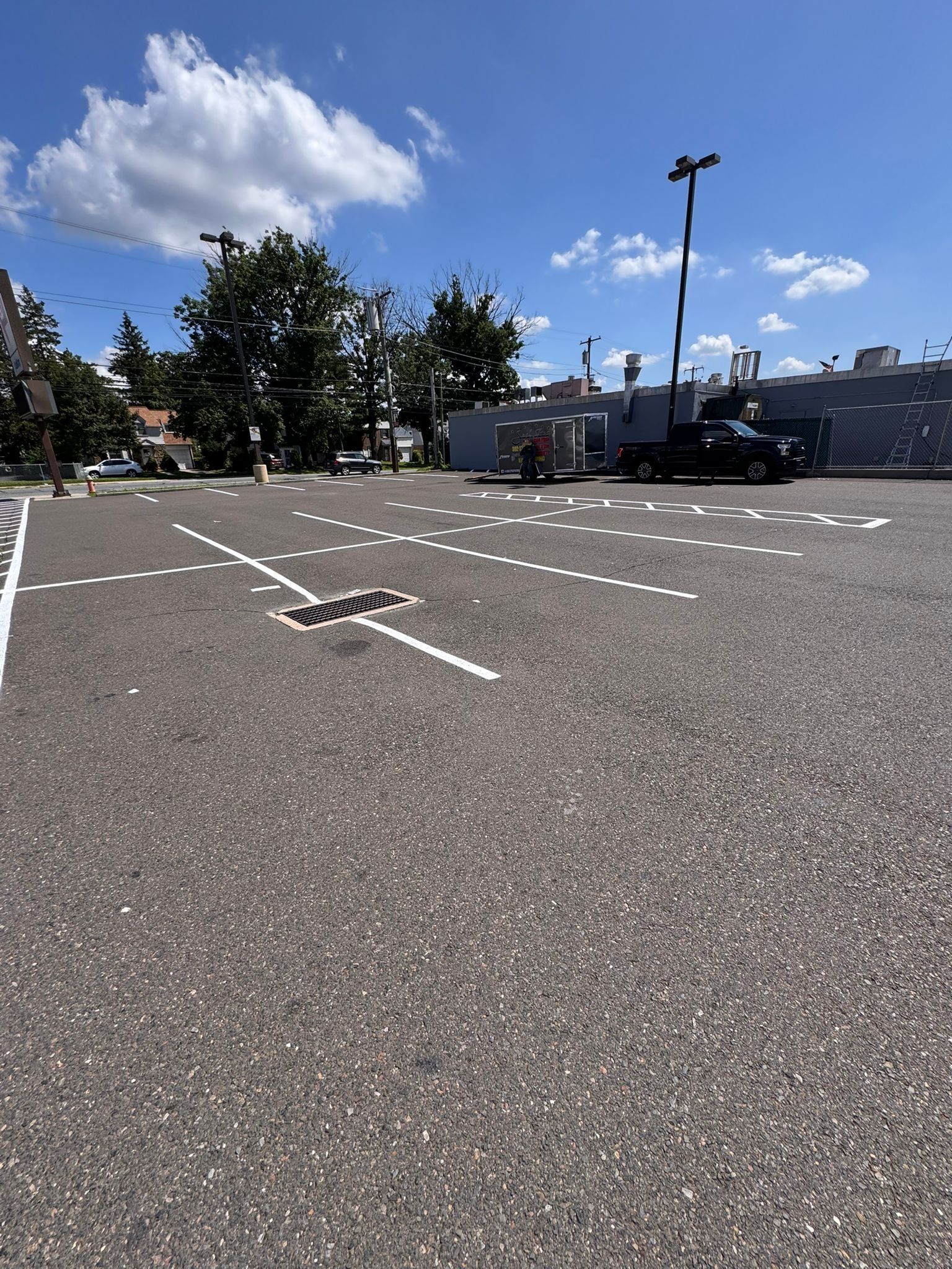 Empty parking lot with white lines and a cloudy blue sky, with trees and buildings in the background