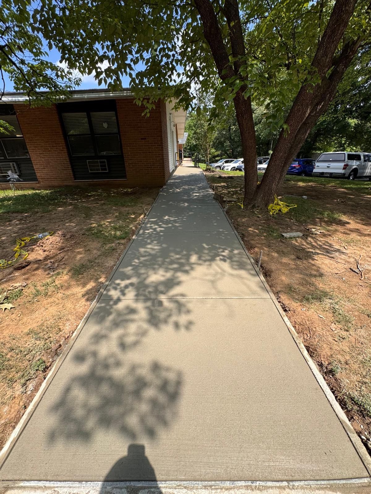 Concrete sidewalk beside a brick building, lined by trees and parked cars in bright sunlight