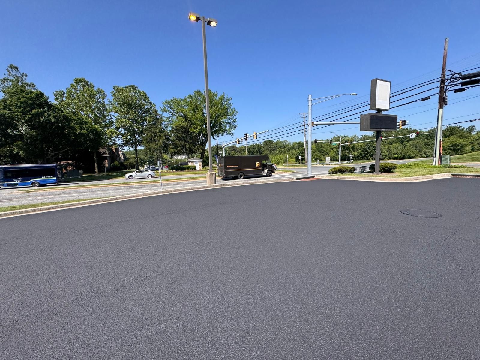Empty parking lot beside a road, with trees, light poles, and utility lines under a clear blue sky.