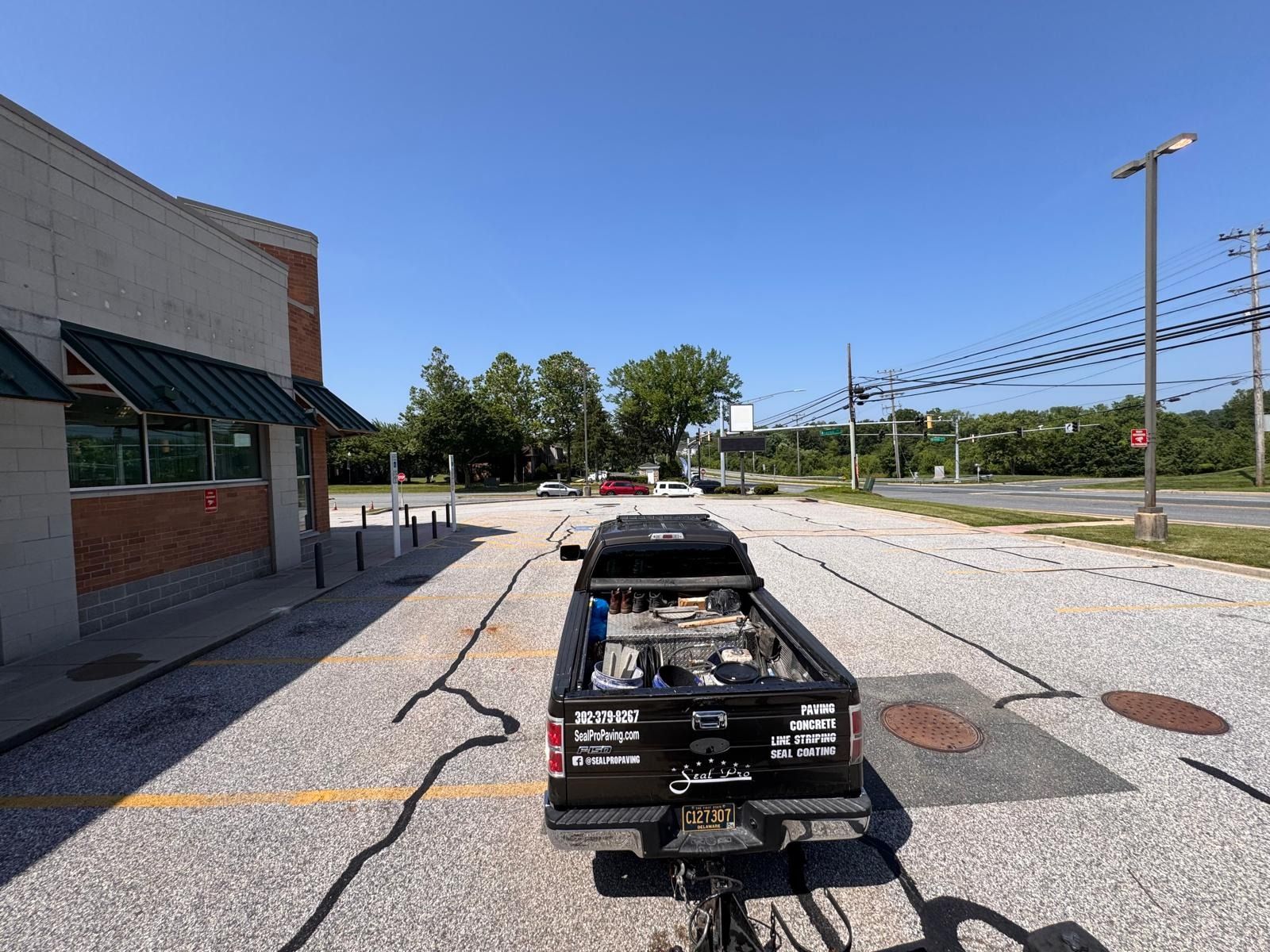 Black utility trailer parked in a gravel lot beside a building under a clear blue sky.